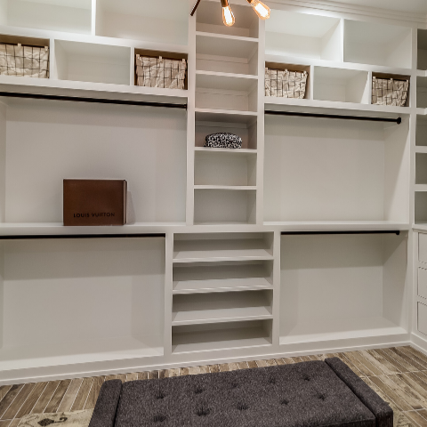 A white built-in walk-in closet system with shelves, hanging rods, a central staircase shelf, drawers on the right, and a gray tufted bench on a wood-look floor.