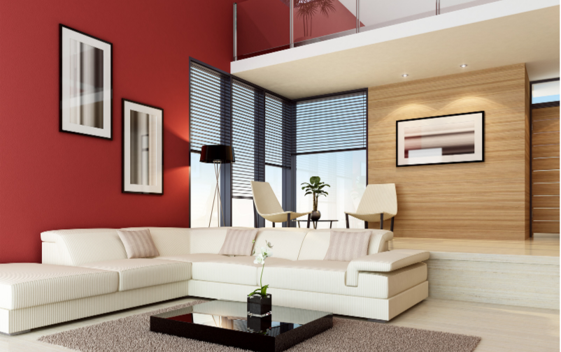 A modern living room interior featuring a large white sectional sofa, a black glossy coffee table, a red accent wall with framed art, wood-paneled walls, and tall windows with blinds.