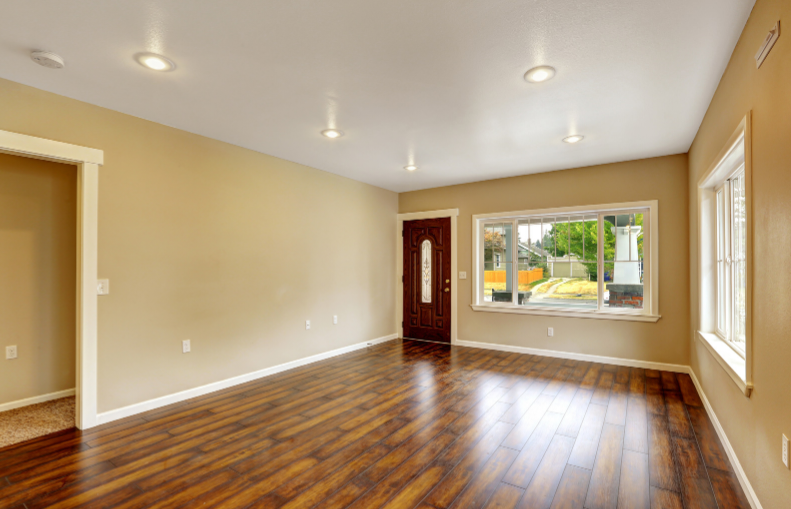 An empty room with beige walls, dark brown wood-look flooring, white baseboards, recessed ceiling lights, and two windows, one of which shows an outdoor view.