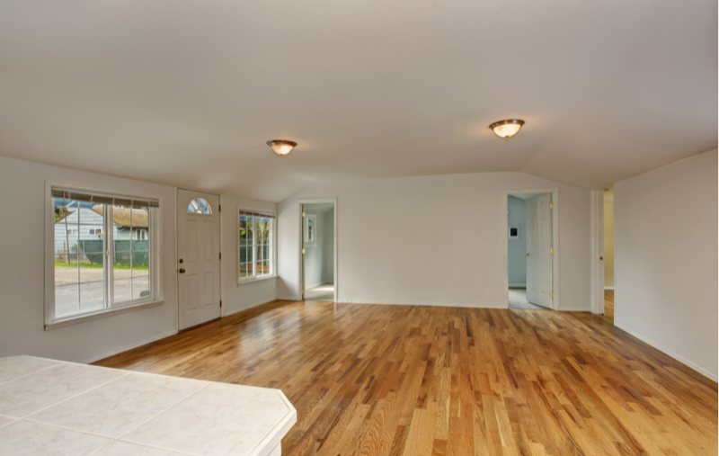 An empty room with white walls, hardwood wood floors, recessed ceiling lights, and multiple windows and doorways.