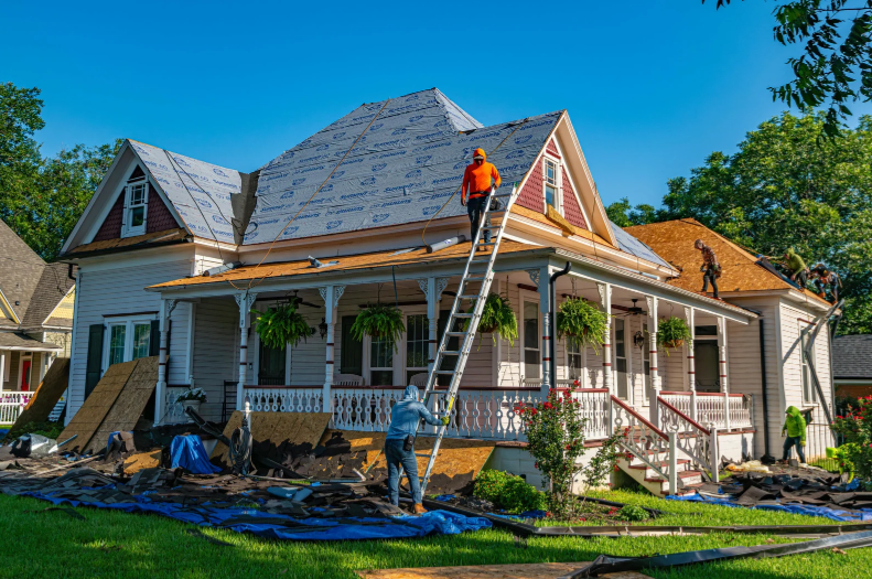 A large white Victorian-style house under construction with workers on the roof and front porch, blue roofing material covering the peak, green trees, and a bright blue sky.