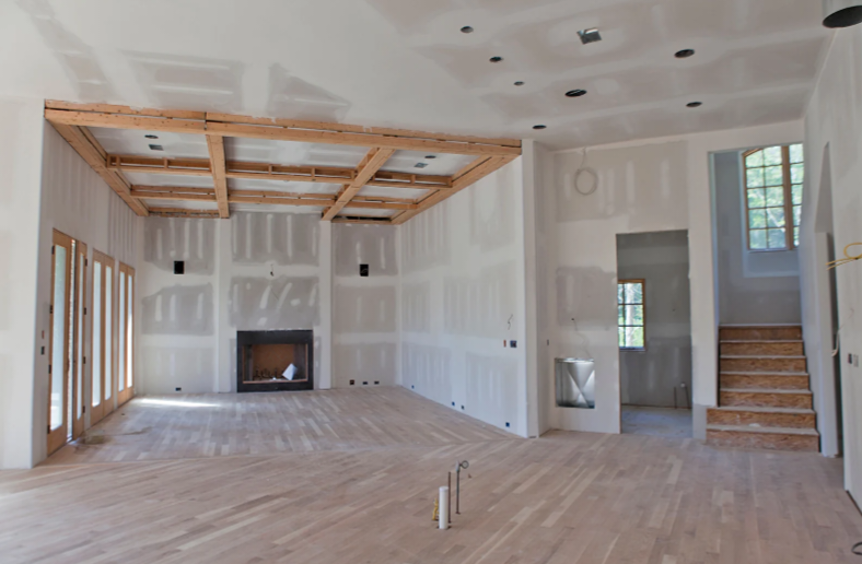 A large, empty room under construction or renovation with white drywall, exposed wooden ceiling joists, light wood flooring, a fireplace on one wall, glass doors on the left, and a staircase on the right.