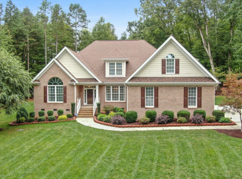 A two-story suburban house with beige siding, brown brick lower walls, brown shutters, a front porch with steps, a manicured green lawn, landscaped shrubs, and a backdrop of tall trees under a blue sky.
