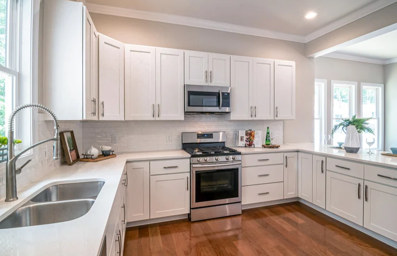 A modern kitchen featuring white shaker-style cabinets, stainless steel appliances including a gas range and microwave, a double sink, hardwood floors, and bright windows.